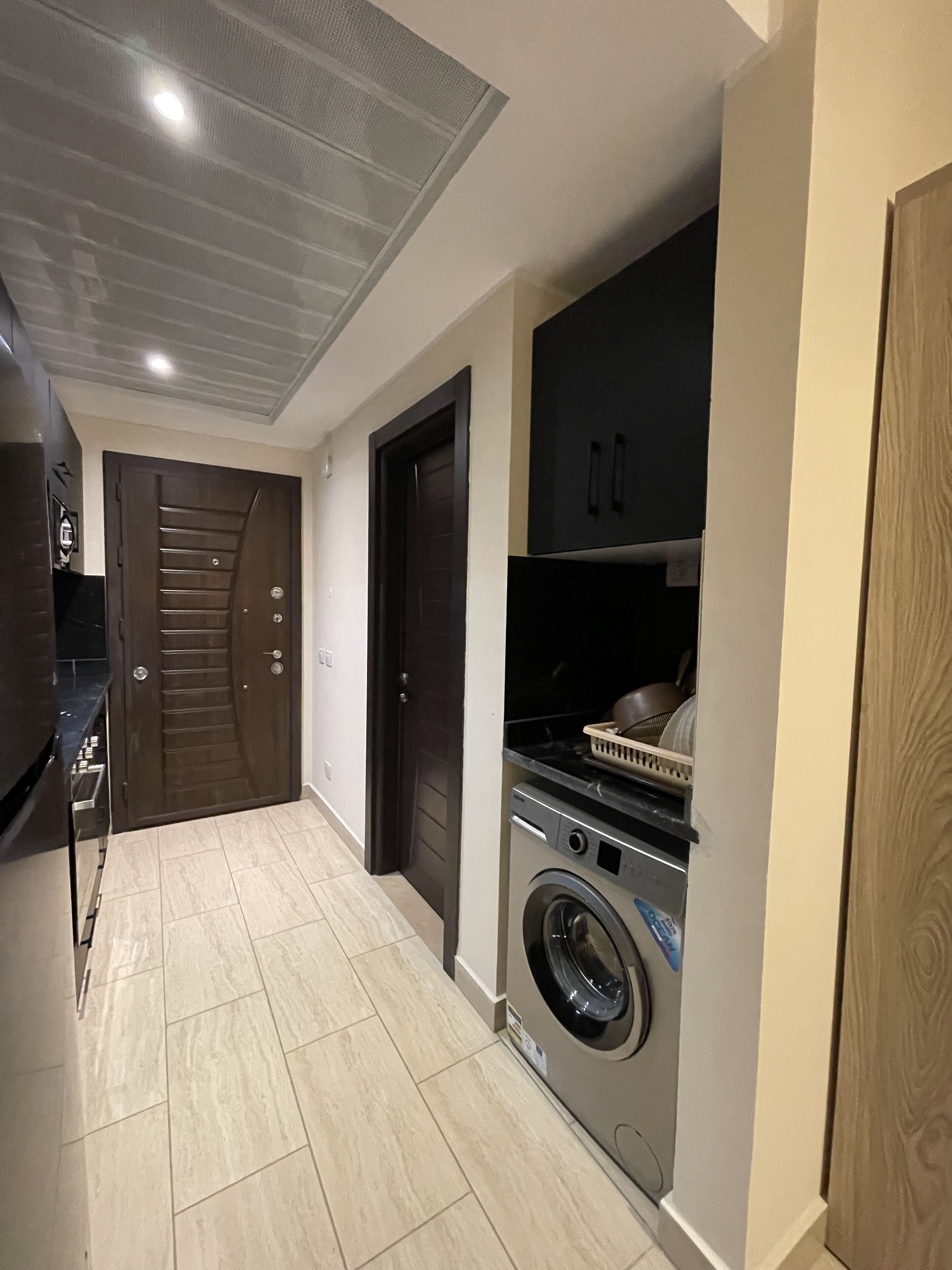 A compact kitchen with a washing machine, a dark wooden door, and a tiled floor.