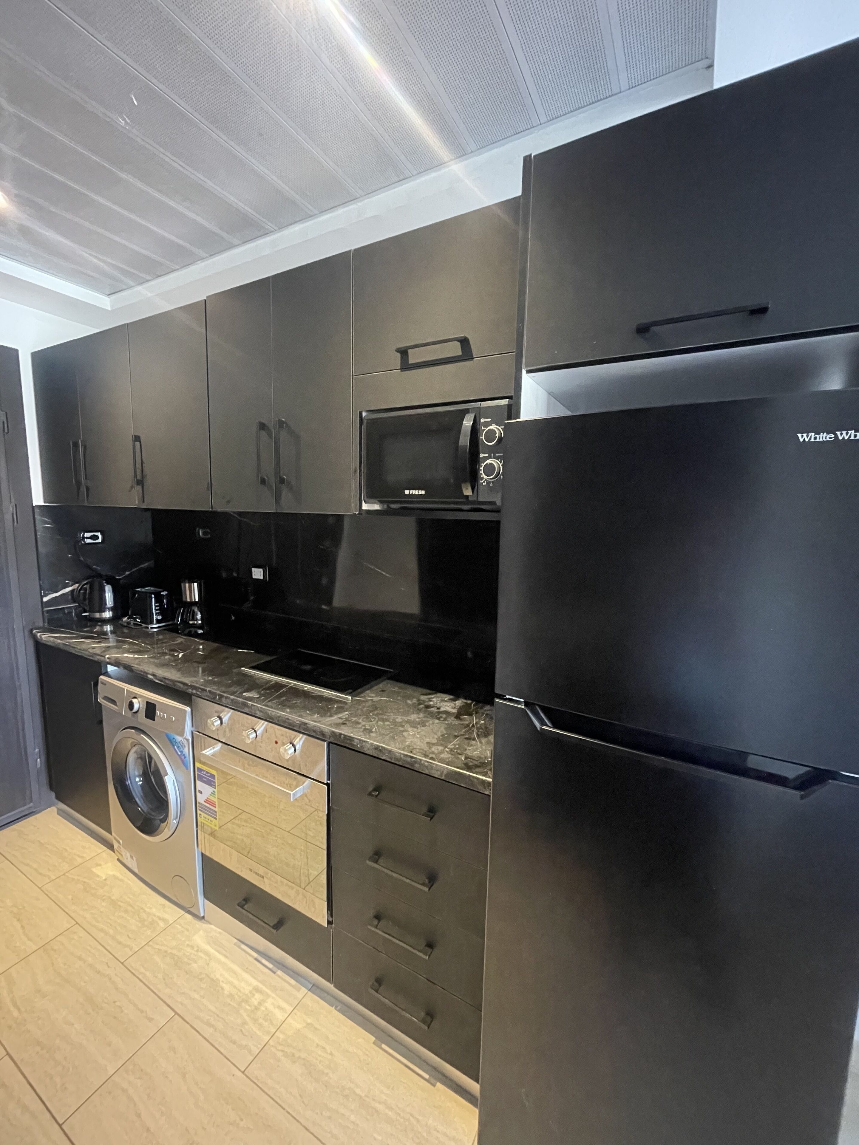 A modern kitchen with black cabinetry, a built-in microwave, a stainless steel refrigerator, a washing machine, and a coffee maker.