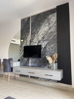 Un salon moderne doté d’un grand téléviseur à écran plat fixé sur un mur en marbre, d’une console grise avec des tiroirs et de deux fauteuils gris.
