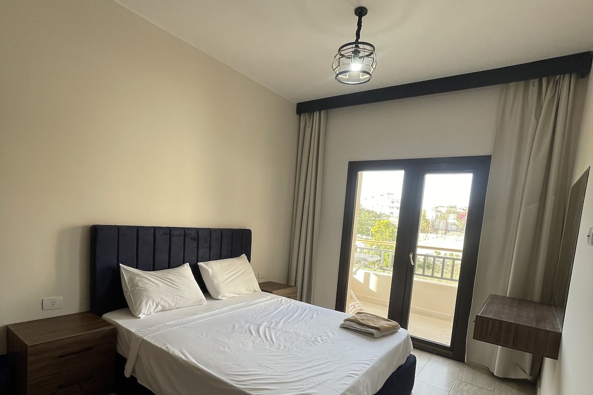 Une chambre à coucher comprenant un lit, des tables de chevet, un ventilateur de plafond, une fenêtre avec des rideaux et une vue sur l’extérieur.