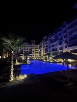 Vista nocturna de un hotel con piscina, palmeras y edificios de varios pisos.