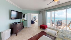 A hotel room with a large TV, a dresser, a red ottoman, a bed, and a balcony with a view of the sea. - Lighthouse 2C - Beautiful Condo at The Lighthouse - Inquire Now for Specials! (Oyster Pond)