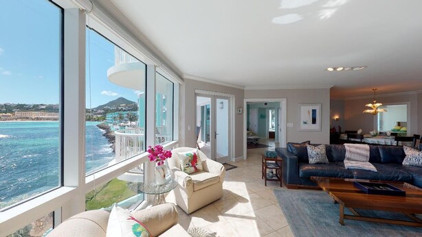 A spacious living room with a large window overlooking the sea, a comfortable sofa, a coffee table, and a dining area with a chandelier. - Lighthouse 2C - Beautiful Condo at The Lighthouse - Inquire Now for Specials! (Oyster Pond)