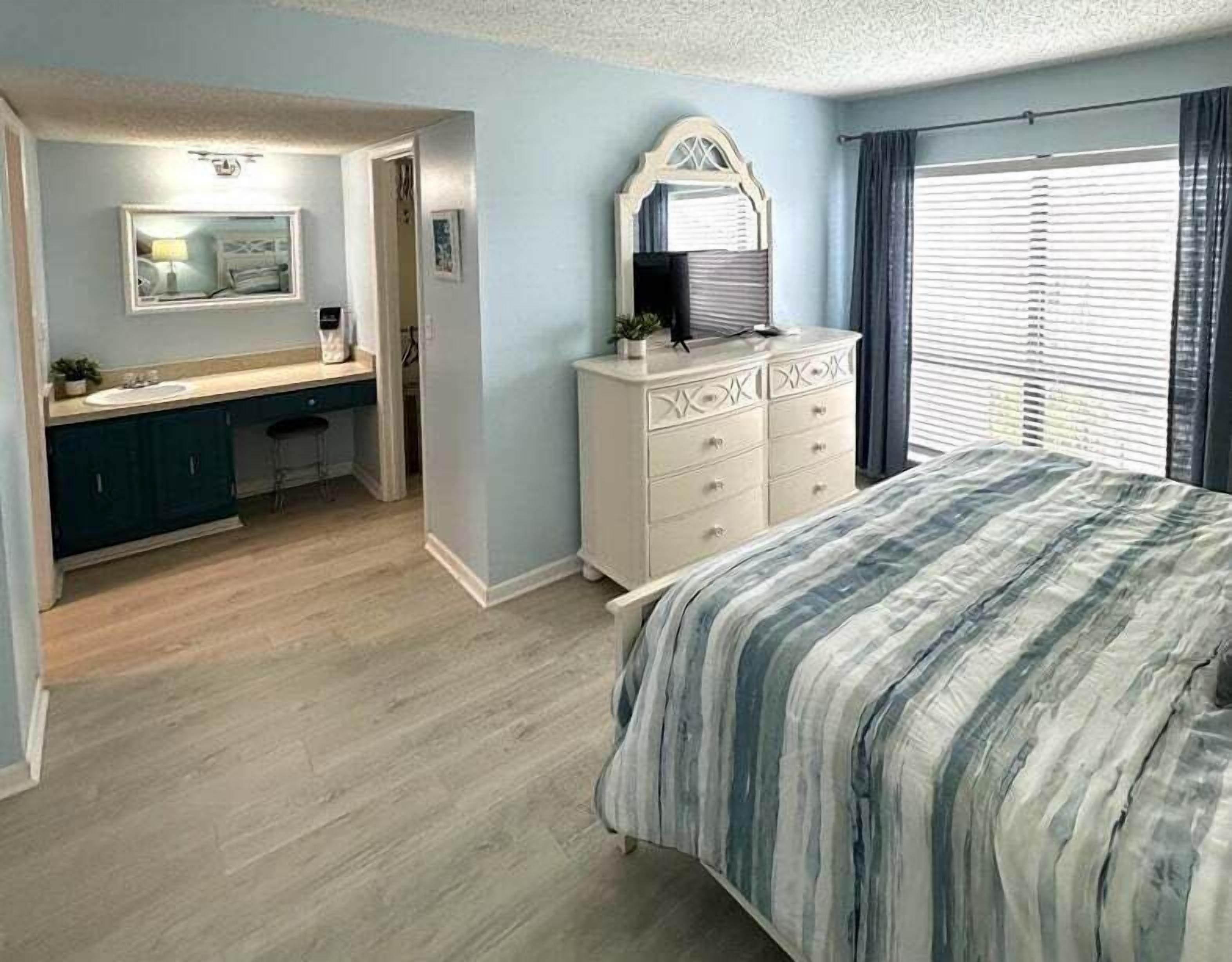 A bedroom with a bed, a dresser, a vanity with a mirror, and a window with curtains.