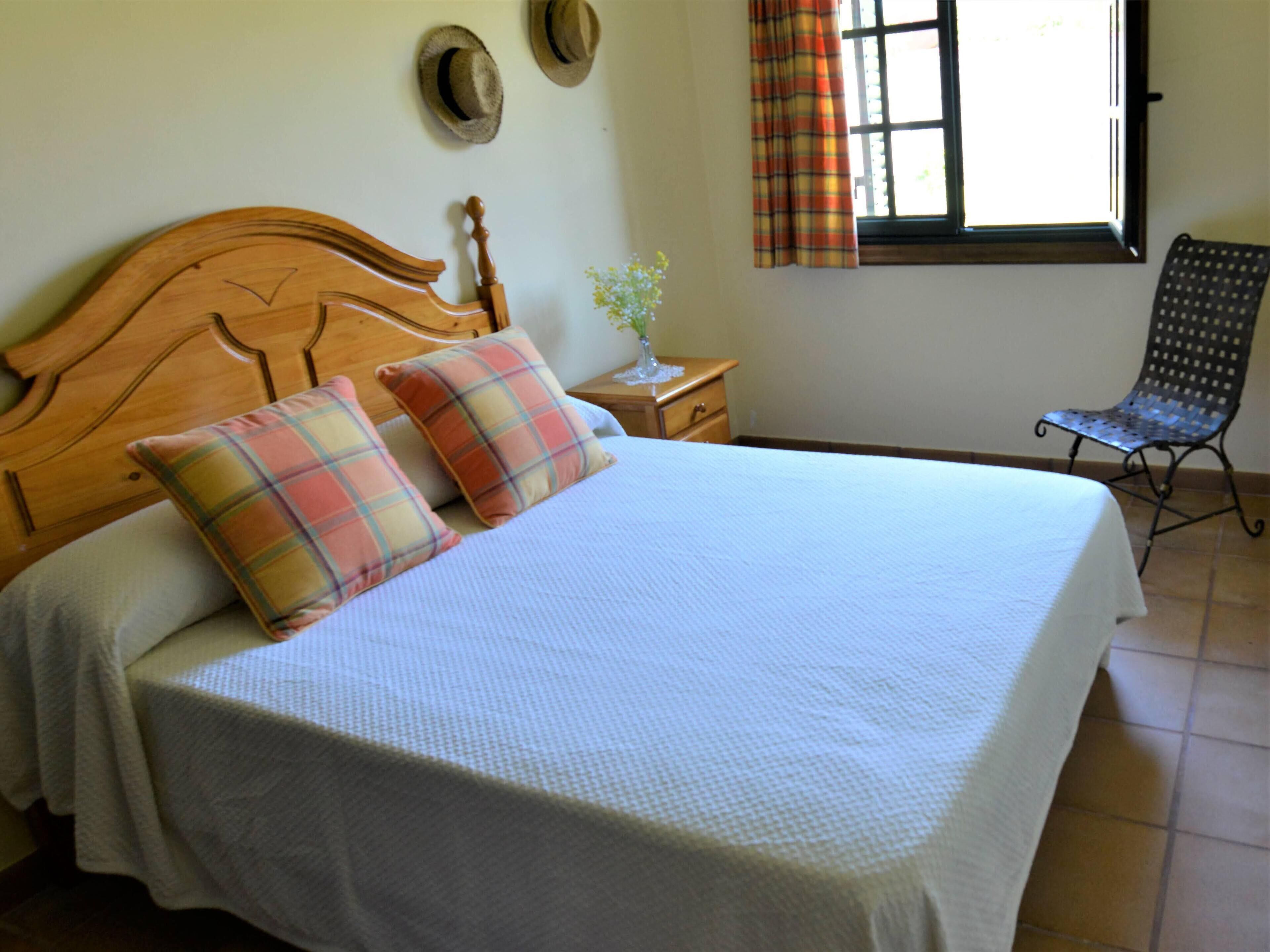 A neatly made bed with plaid pillows, a wooden headboard, a nightstand with a vase, and a chair by a window with curtains.