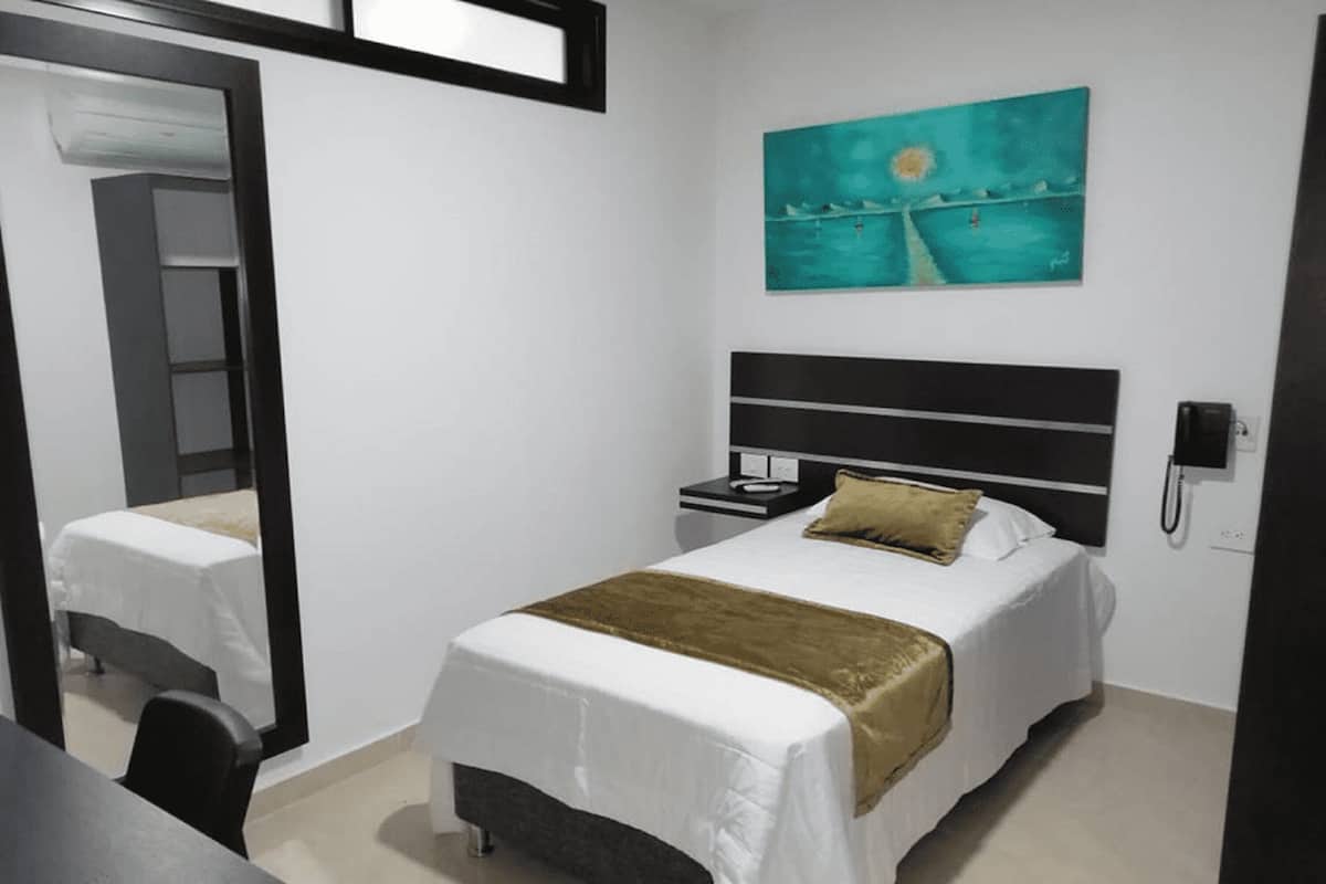 A hotel room with a bed, a nightstand, a bedside table, a mirror, a painting, and a telephone.