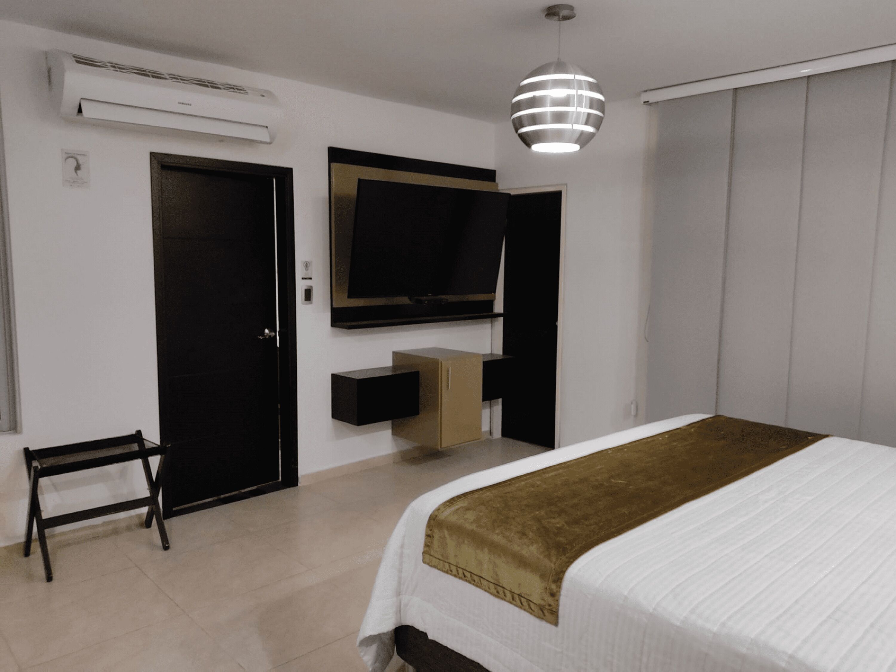 A bedroom with a bed, a TV mounted on the wall, a bedside table, a wardrobe, and a hanging light fixture.