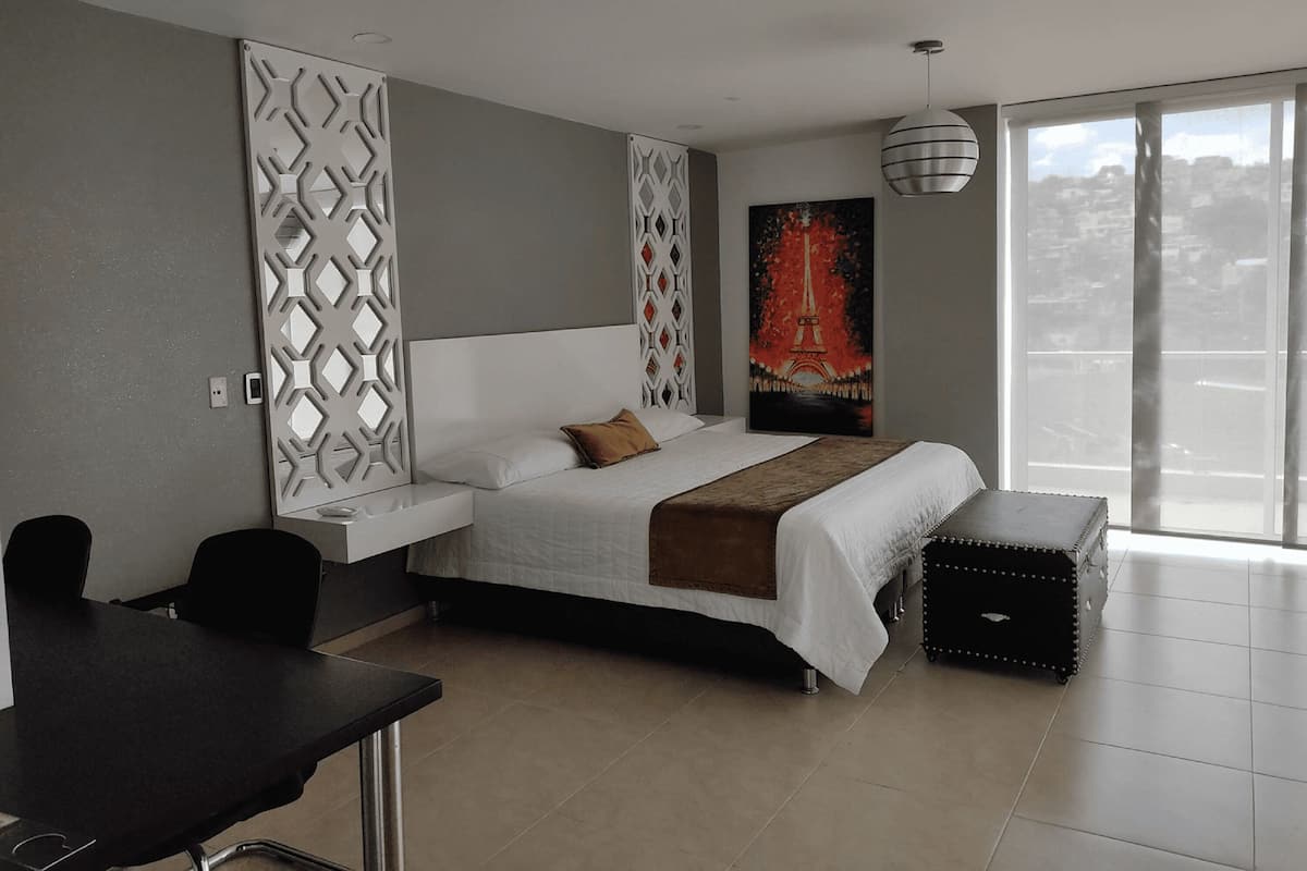 A modern bedroom with a large bed, a desk, and a view of the city through a large window.