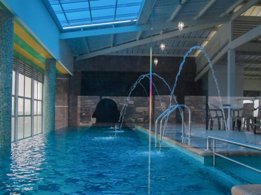 Una piscina interior con agua cristalina, techo de cristal y pared de piedra al fondo.
