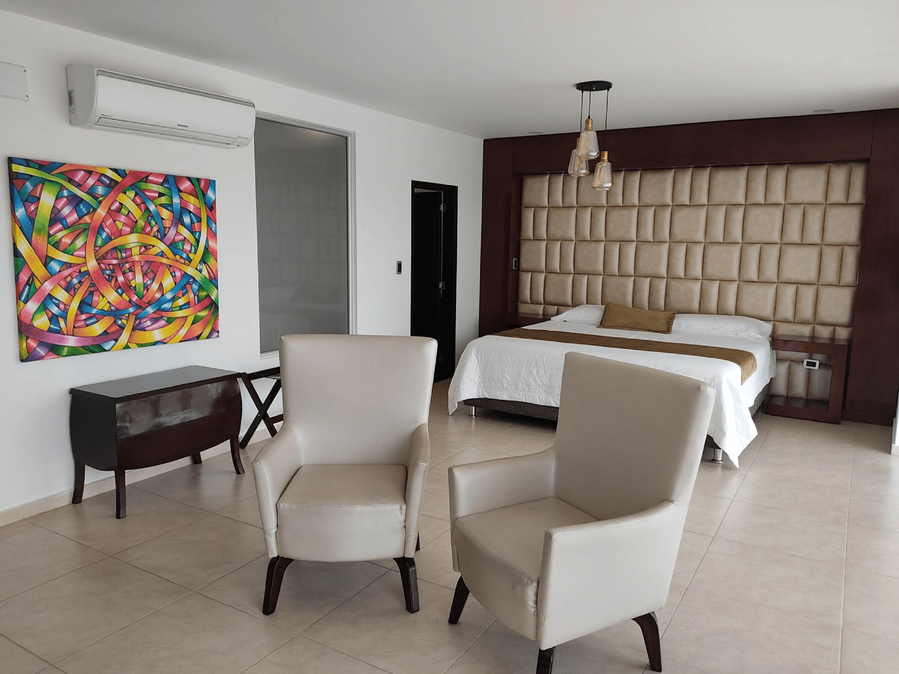 A modern hotel room with a large bed, a headboard with a tufted design, a desk, and a colorful abstract painting on the wall.