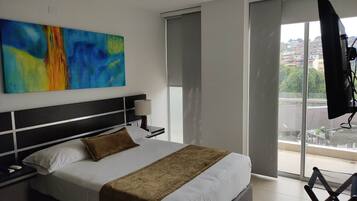 A hotel room with a large bed, a nightstand, a lamp, a colorful abstract painting, and a view of the city through a large window.