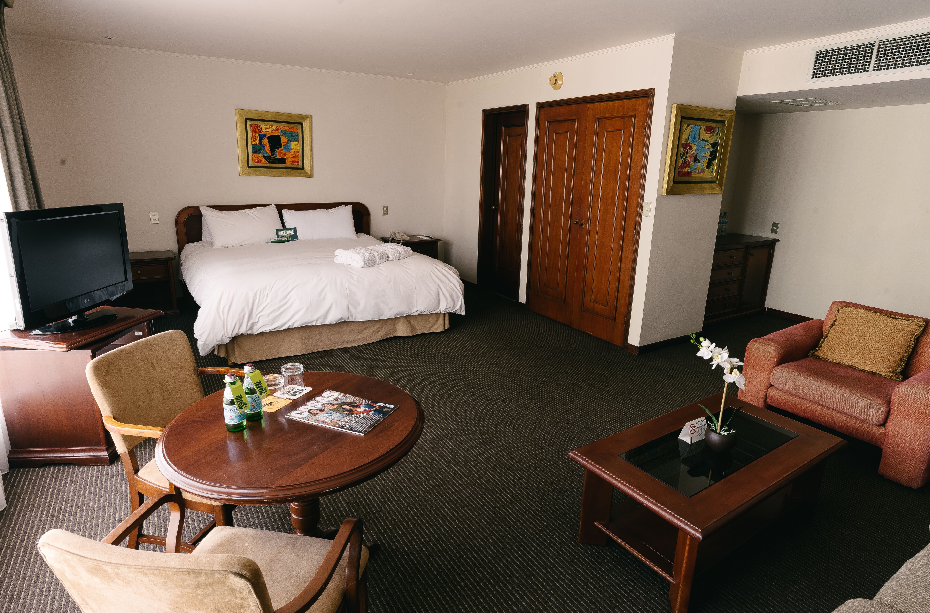 A hotel room with a bed, a TV, a dining table with chairs, a sofa, and two doors.