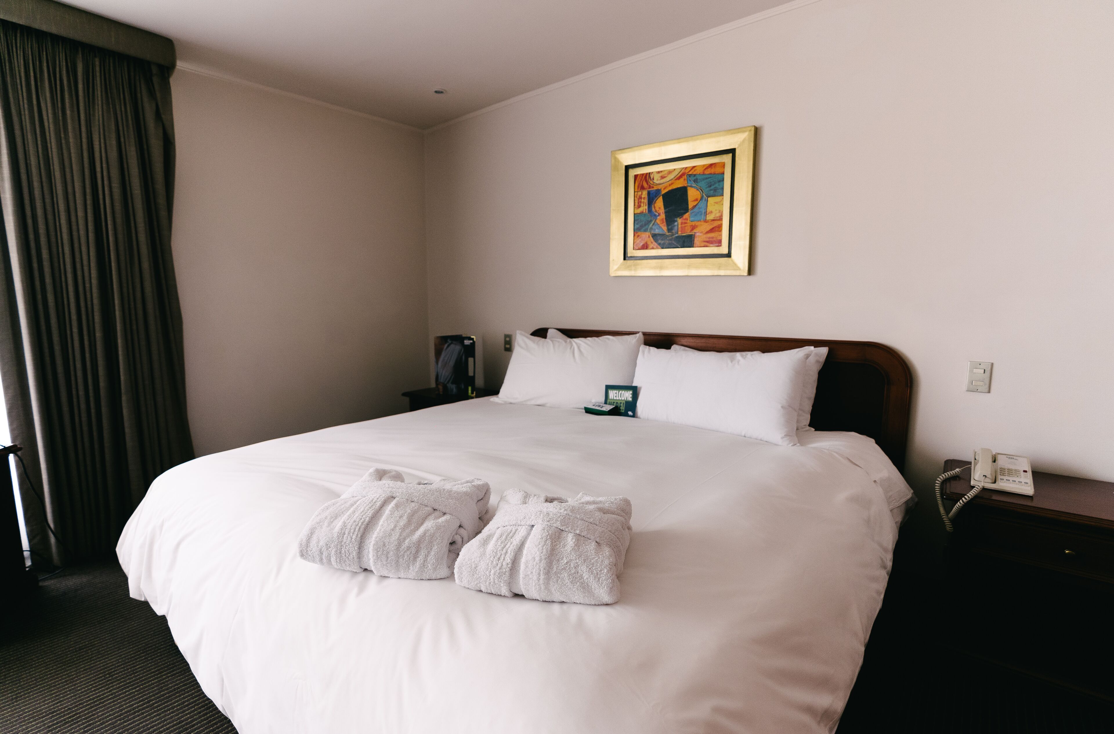A hotel room with a large bed, white linens, two towels, a painting on the wall, a bedside table with a phone, and a window with curtains.