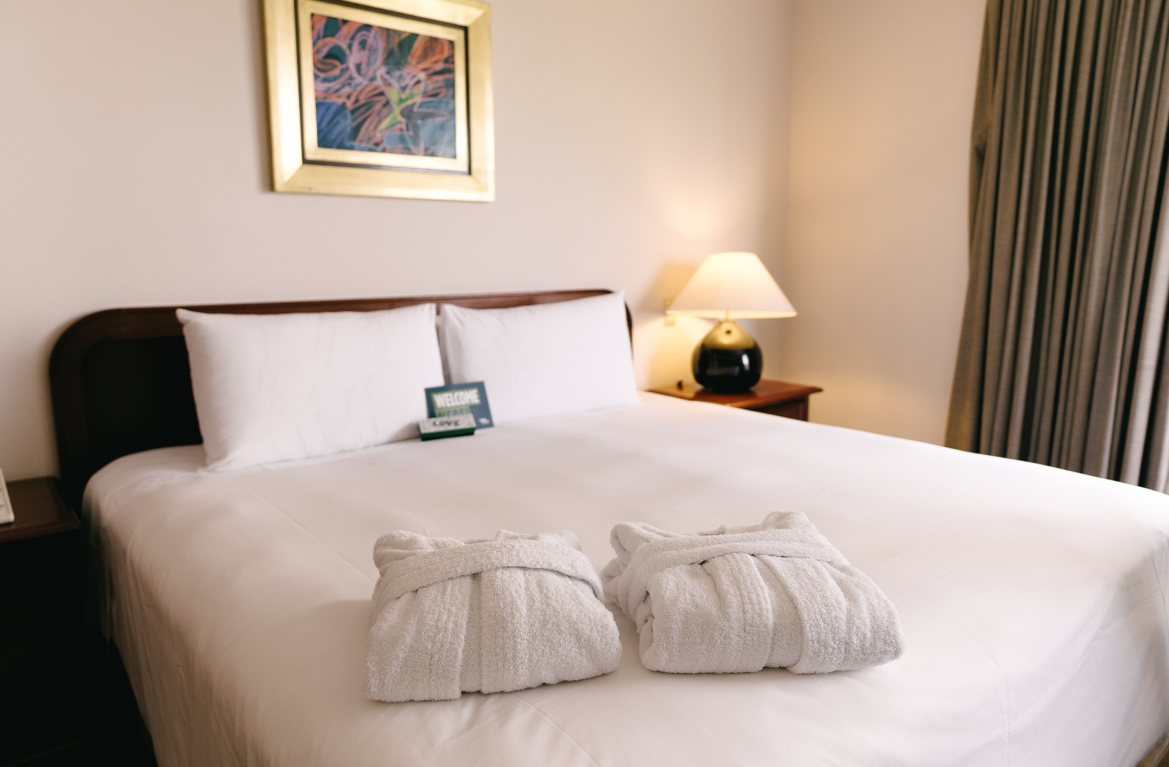 Habitación de hotel con una cama bien hecha, una lámpara de noche, un cartel de bienvenida y una obra de arte enmarcada en la pared.