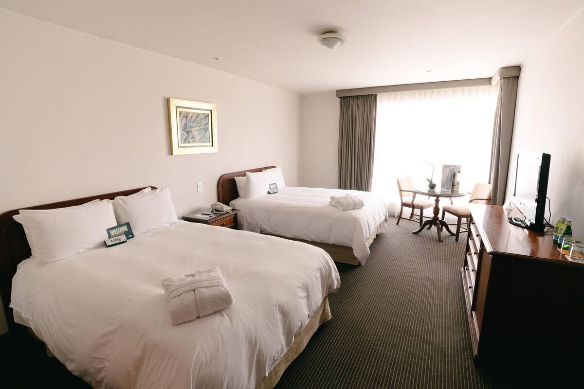 Habitación de hotel con dos camas, un escritorio, una silla, una mesita y una ventana con cortinas.