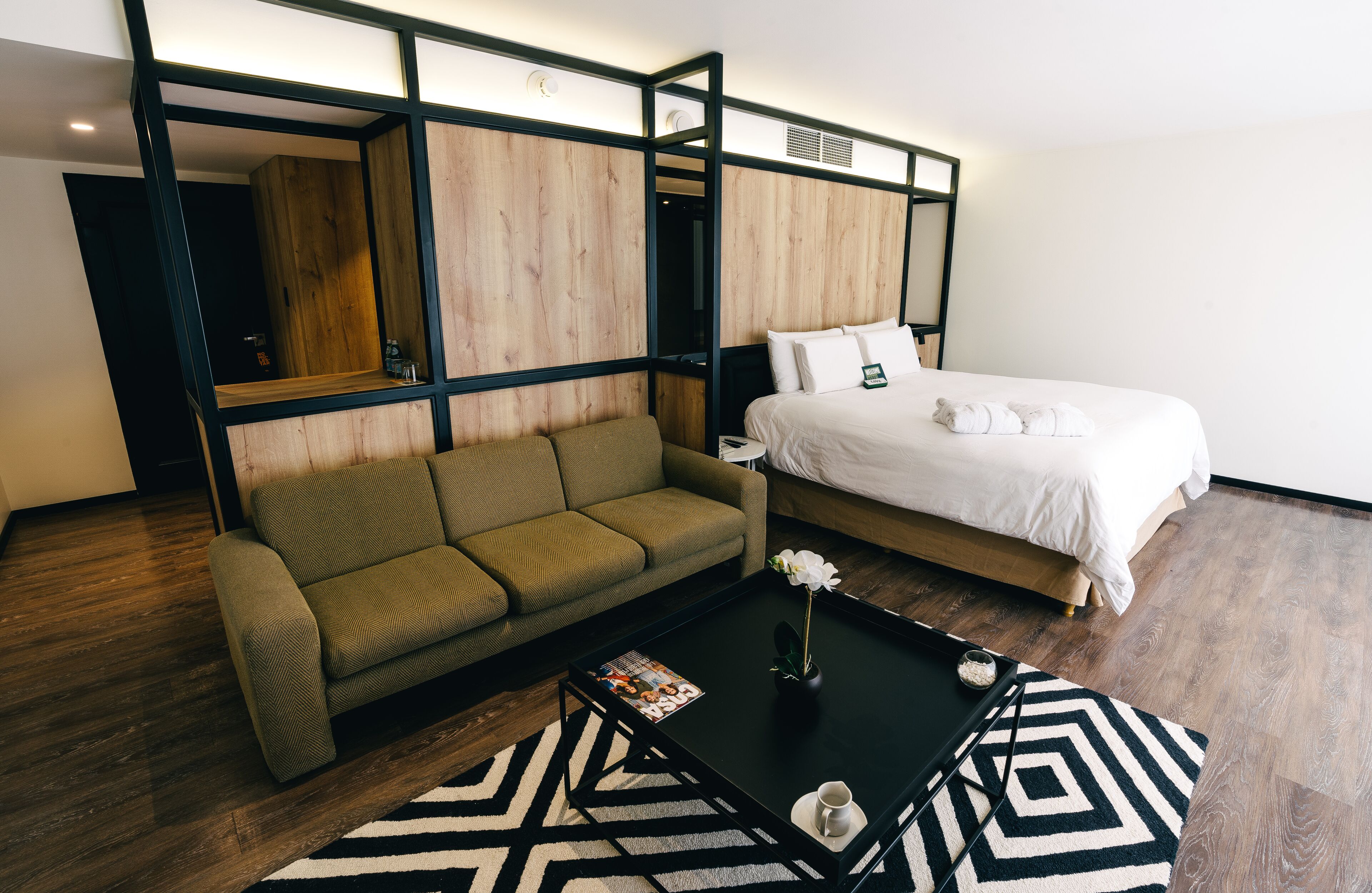 A modern hotel room with a wooden partition, a green sofa, a bed with white linens, and a coffee table with a magazine and a flower vase.