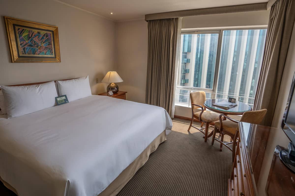 Habitación de hotel con una cama grande, una mesita con sillas, una lámpara y un ventanal con cortinas.
