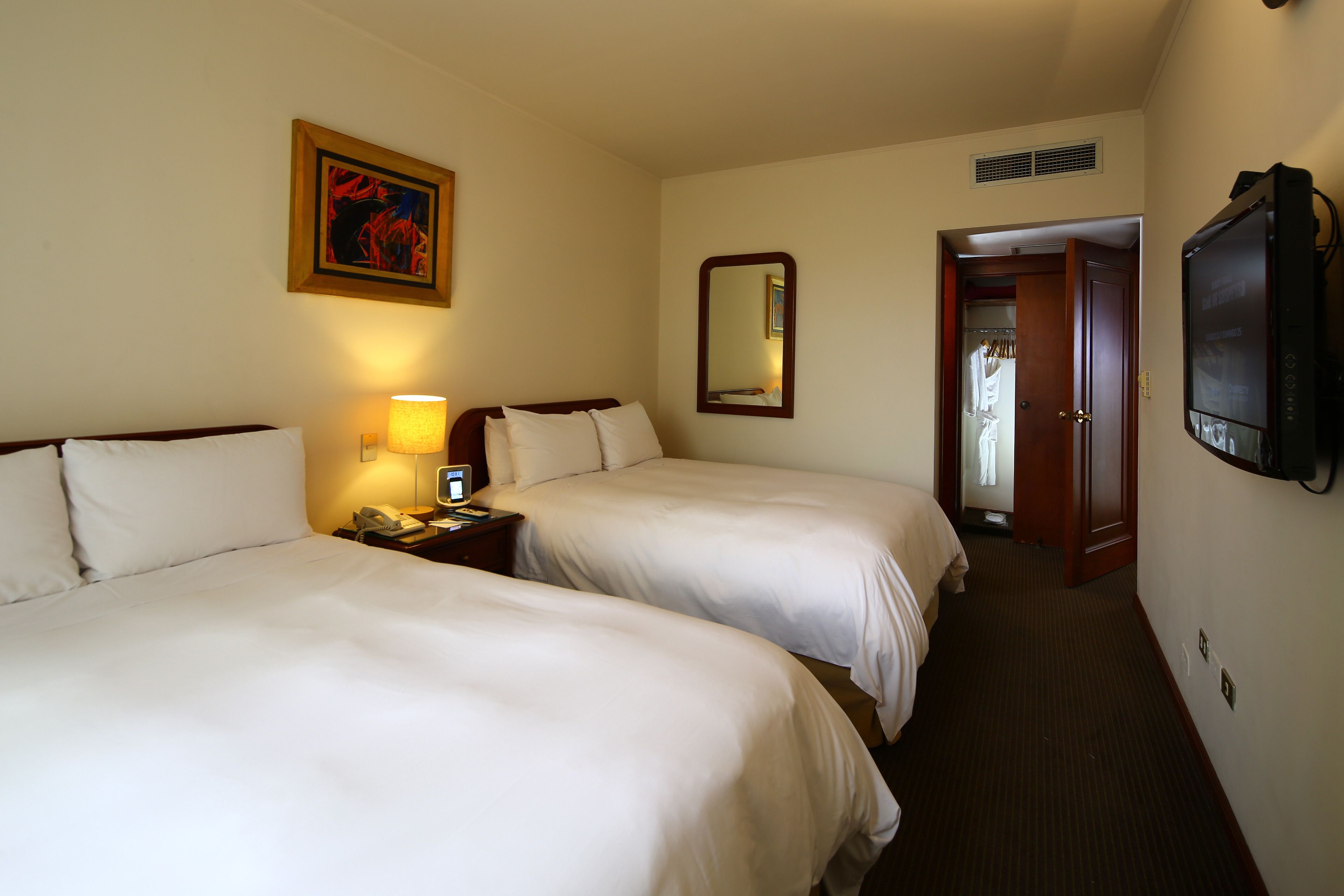 Une chambre d’hôtel avec deux lits, une table de chevet avec une lampe, une télévision, un miroir et une porte donnant sur un placard.