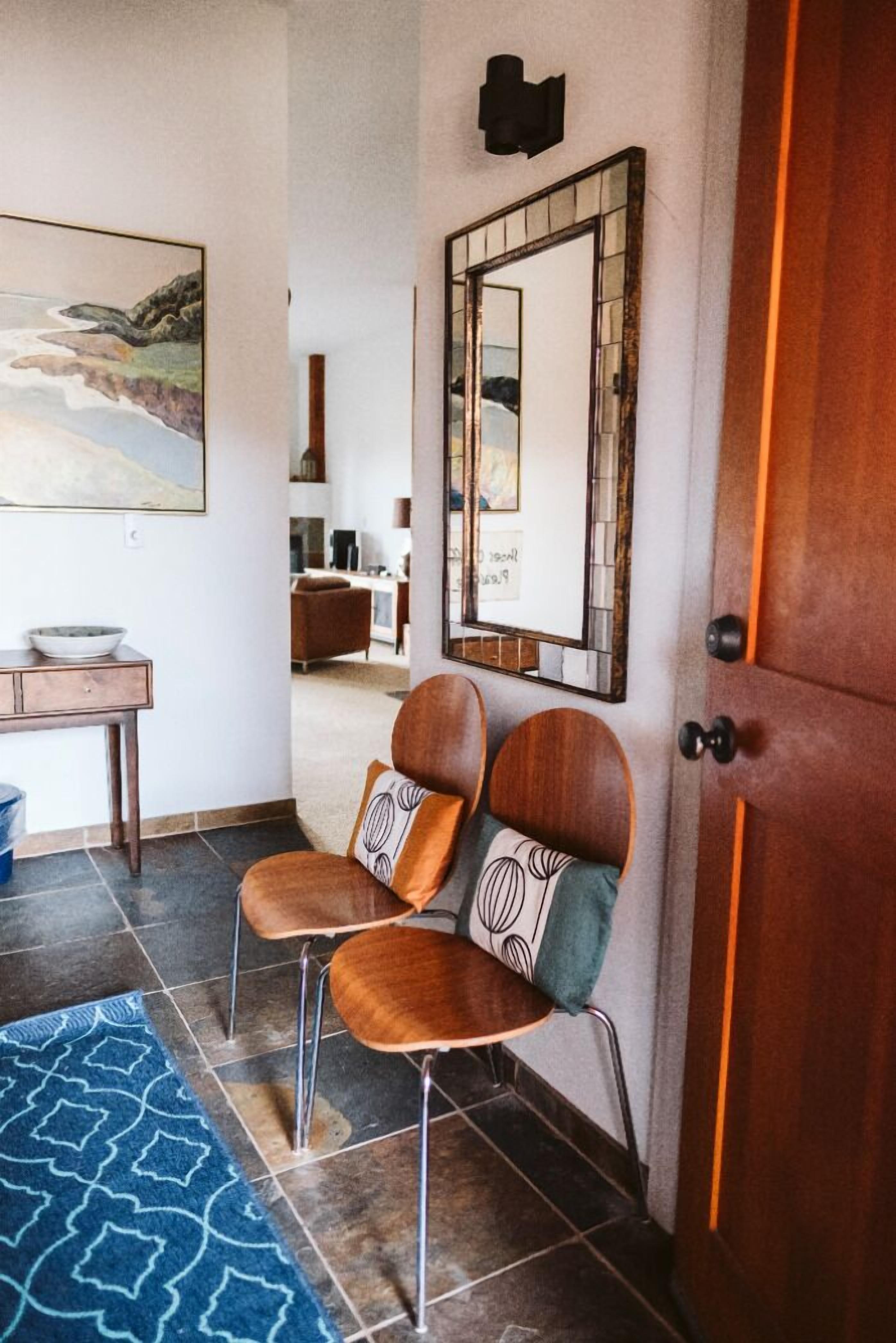 Un pasillo con una puerta de madera, un espejo y una zona de estar con dos sillones.