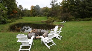 A pond with white Adirondack chairs arranged around it, surrounded by a grassy area and trees. - Vermont Farmhouse in the Heart of the Green Mountains (Granville)