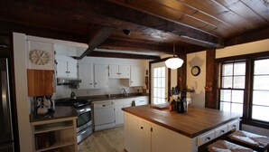A kitchen with wooden beams, white cabinets, a central island with stools, and a large window. - Vermont Farmhouse in the Heart of the Green Mountains (Granville)