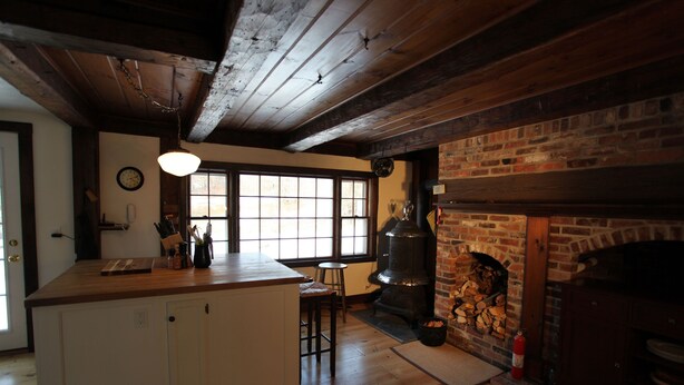 A kitchen with wooden beams, a brick fireplace, and a large window. - Vermont Farmhouse in the Heart of the Green Mountains (Granville)