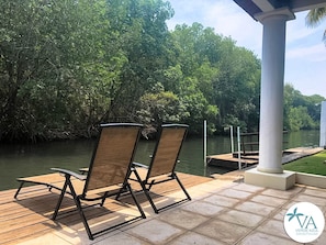 Dos sillones de descanso en una terraza de madera con vistas a un cuerpo de agua, con un pilar blanco y un muelle al fondo.