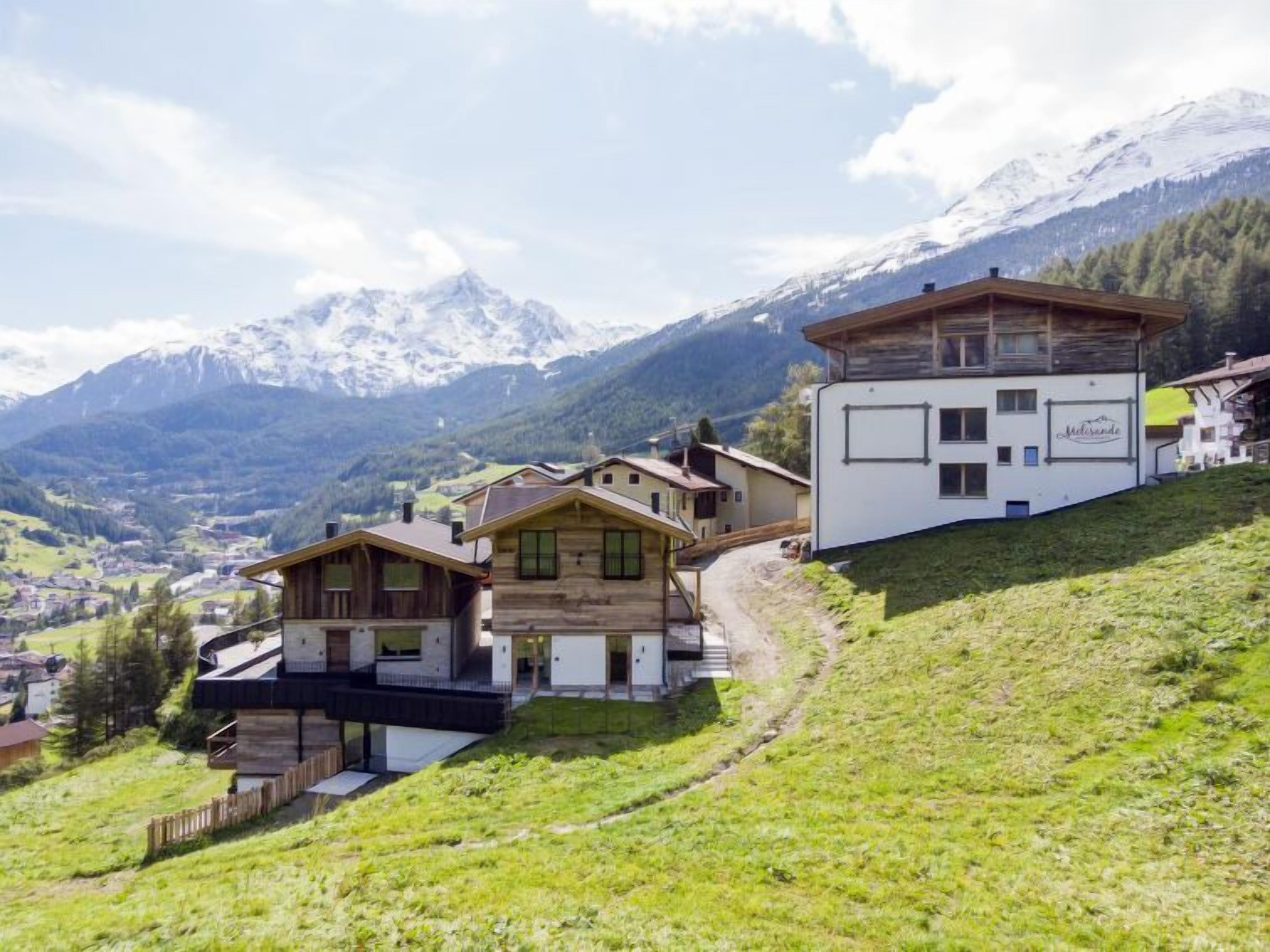 Un albergue de montaña con cabañas de madera y un edificio blanco con un cartel, rodeado de montañas nevadas y un valle abajo.