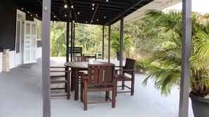 An outdoor patio with a wooden table and chairs, surrounded by greenery and a covered area with a metal roof.