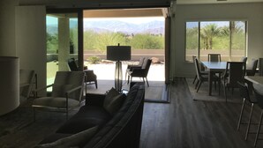 Ein modernes Wohnzimmer mit einer Couch, Sesseln, einem Couchtisch und einem Essbereich mit Tisch und Stühlen. Große Fenster bieten Ausblick auf Berge und Grünflächen.