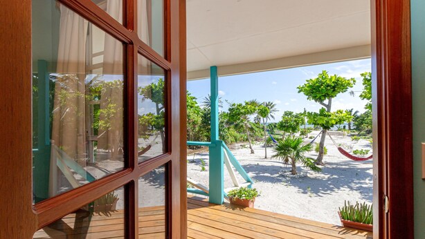 Una terraza de madera con plantas en macetas, una hamaca y vistas a una playa de arena con palmeras.
