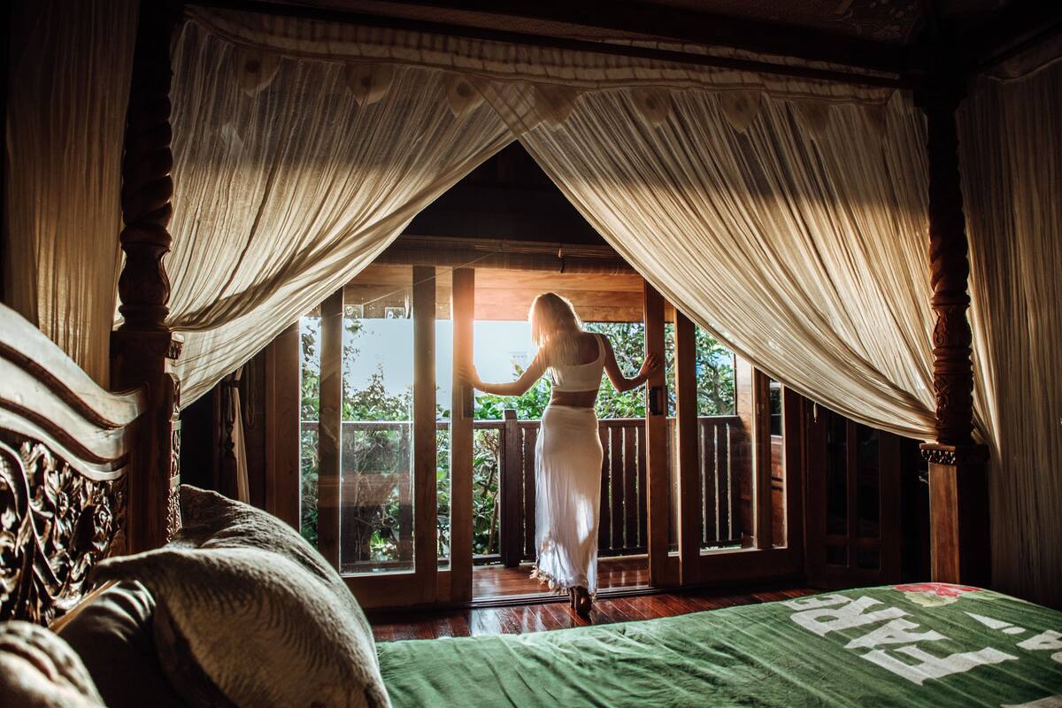 A room with a bed, a window with curtains, and a view of greenery outside.