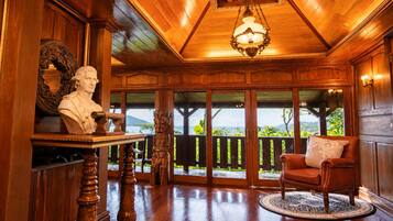 A room with a wooden ceiling, a chandelier, a bust on a table, a sofa, and a view of the outdoors.