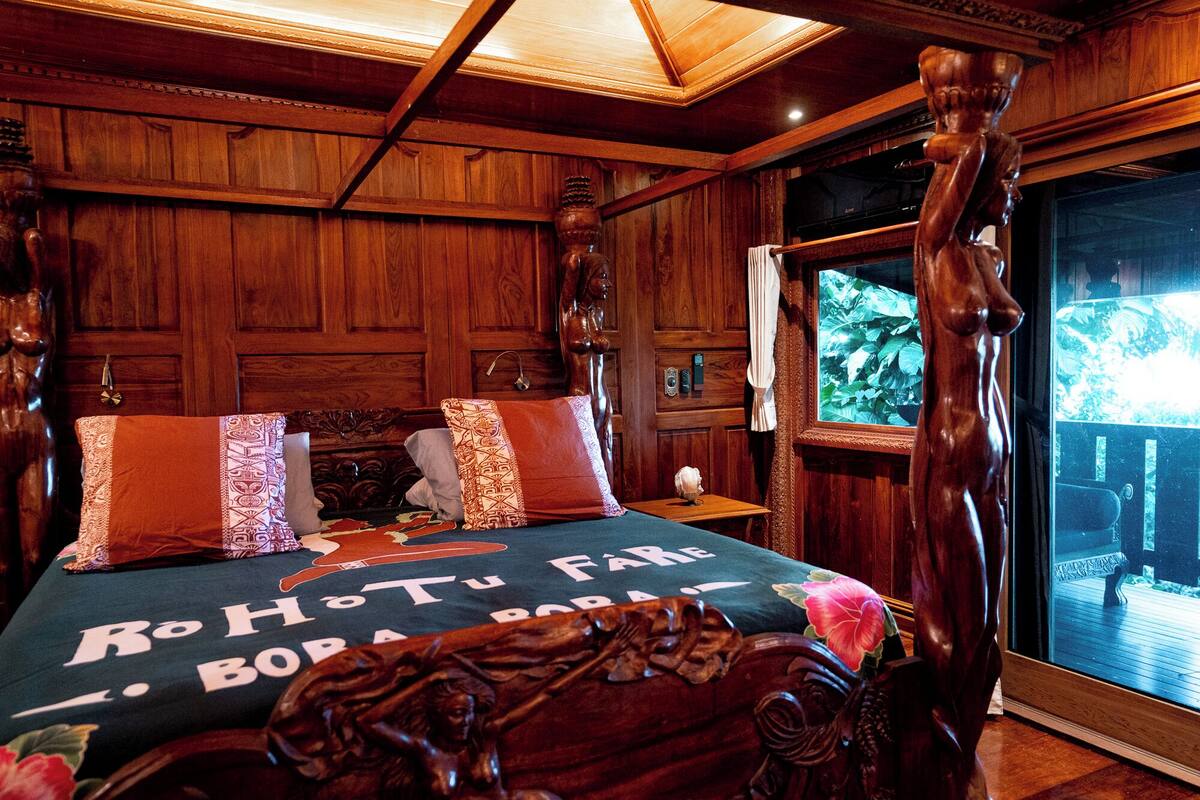 A wooden four-poster bed with a floral bedspread, a wooden headboard, and a view of greenery through a window.