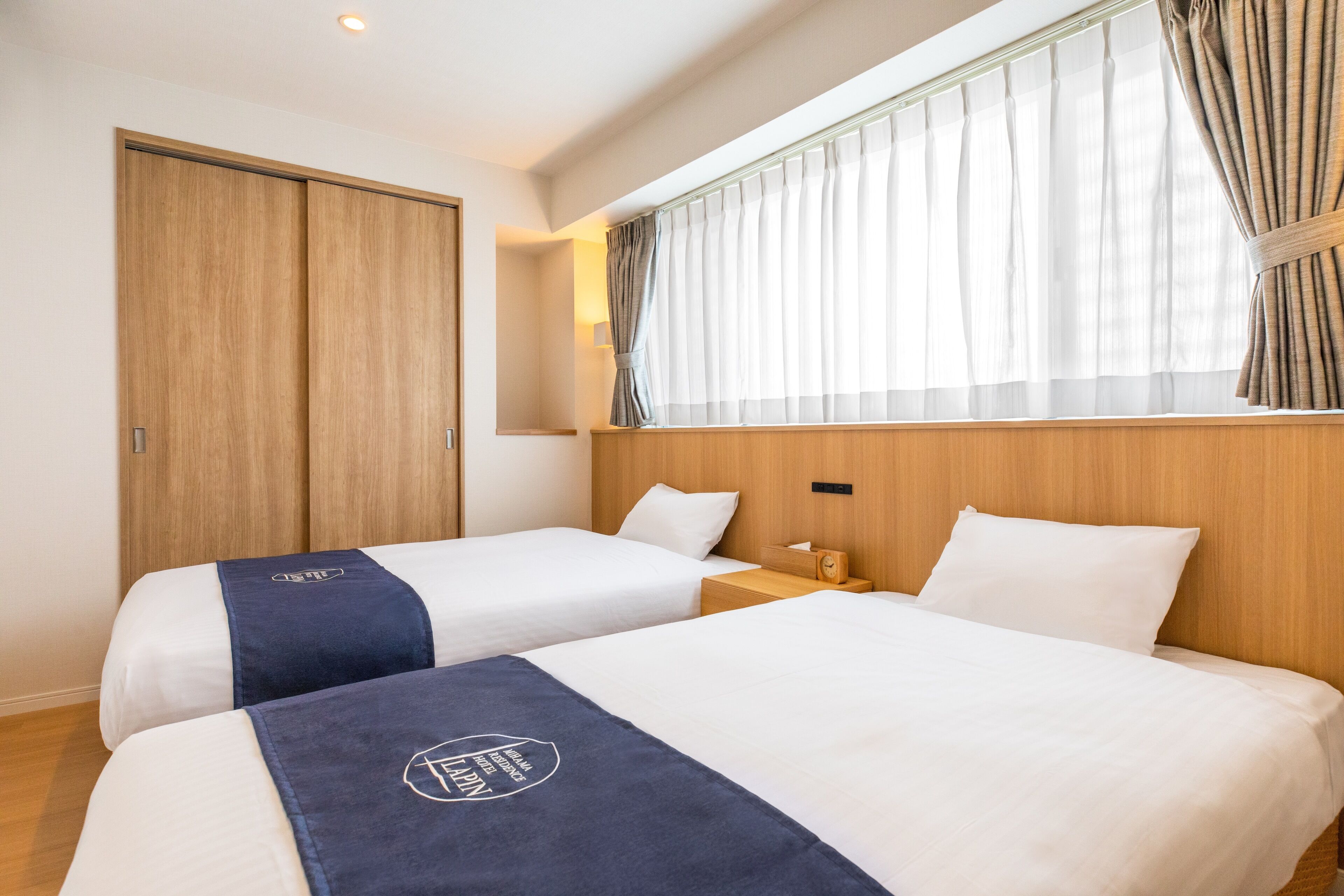 A hotel room with two beds, a wooden wardrobe, a window with curtains, and a nightstand with a lamp.