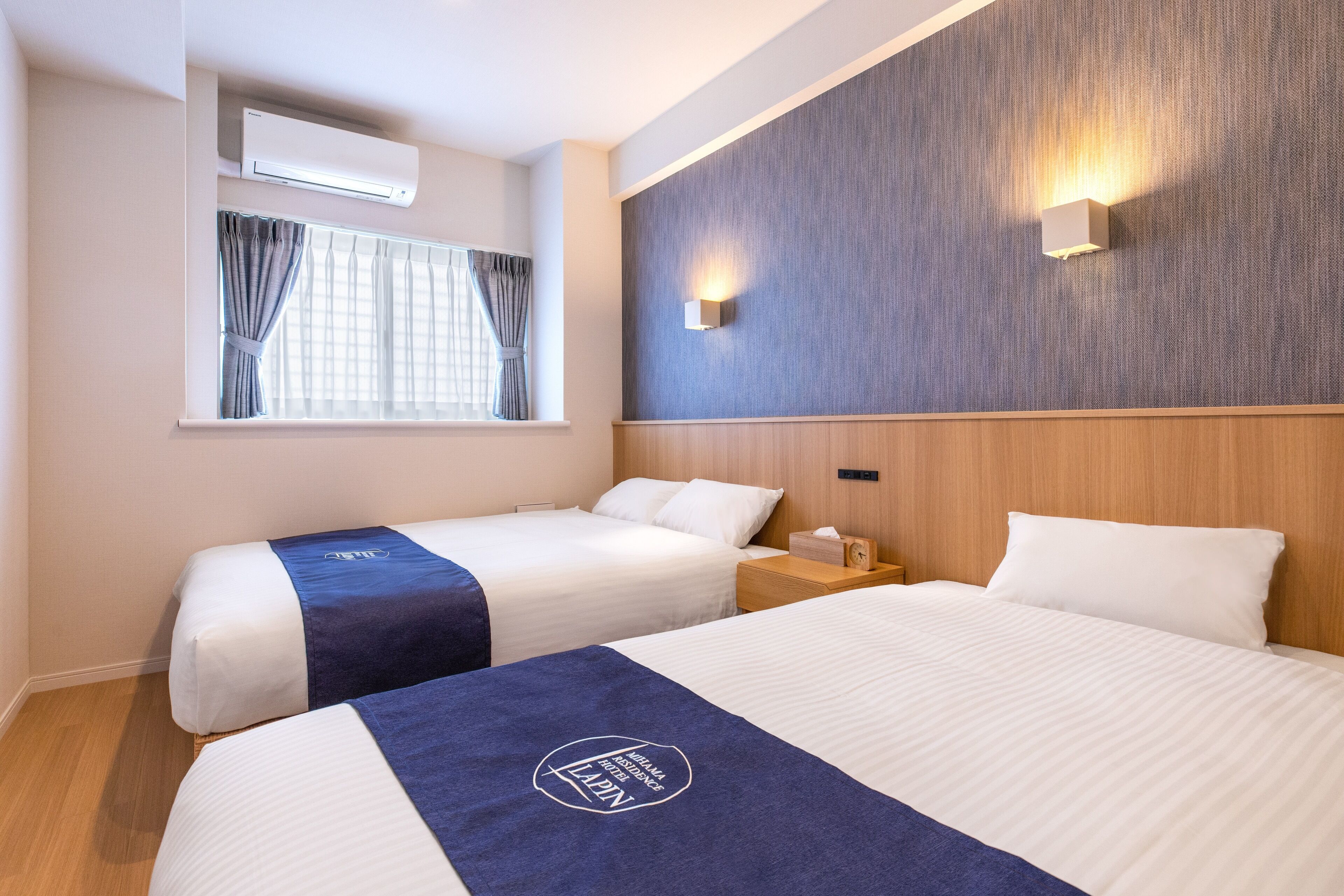 Habitación de hotel con dos camas, una ventana con cortinas, un equipo de aire acondicionado, una mesita de noche y una lámpara fijada a la pared.