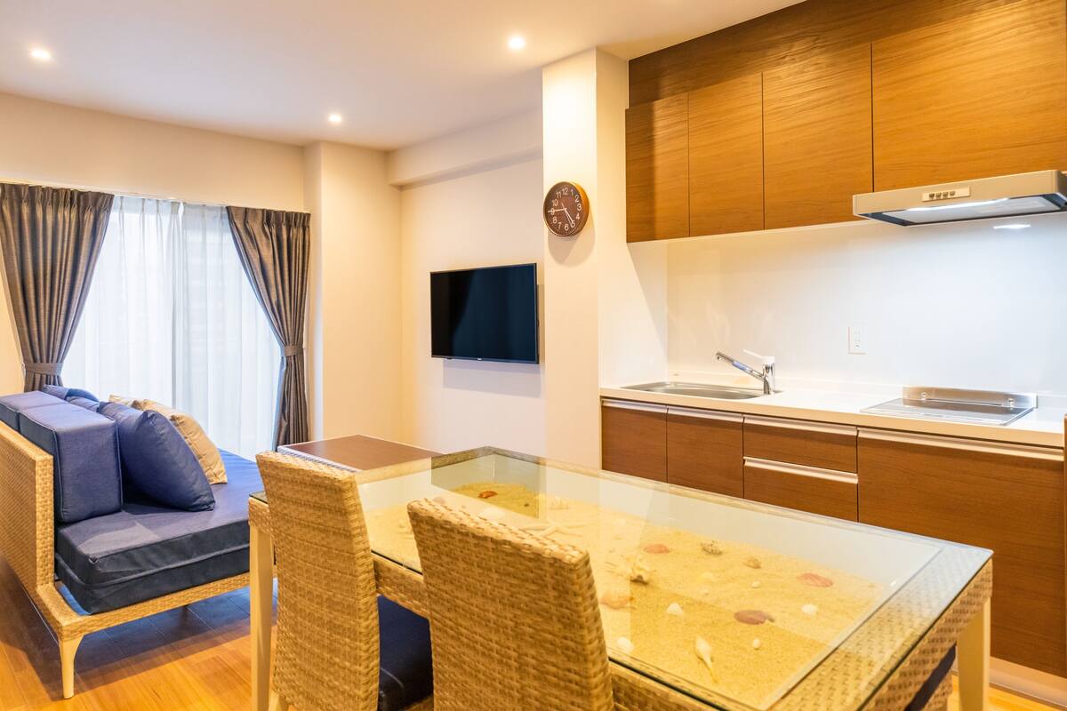 Uma sala de estar moderna com sofá, mesa de jantar de vidro, cadeiras de vime, cozinha com armários de madeira, forno embutido e televisão de tela plana.
