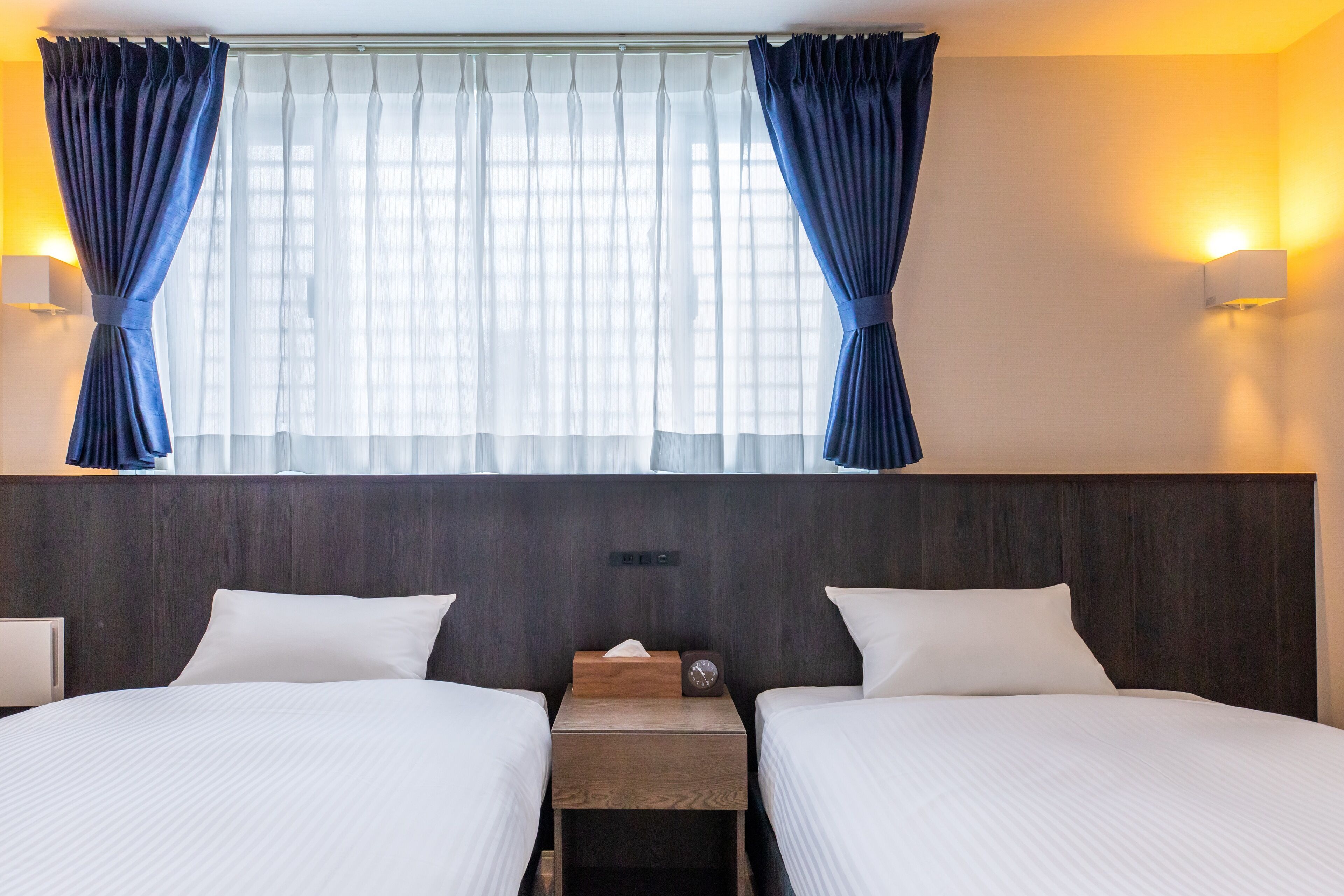 Een hotelkamer met twee bedden, een houten hoofdeinde, een nachtkastje met een klok en een raam met gordijnen.