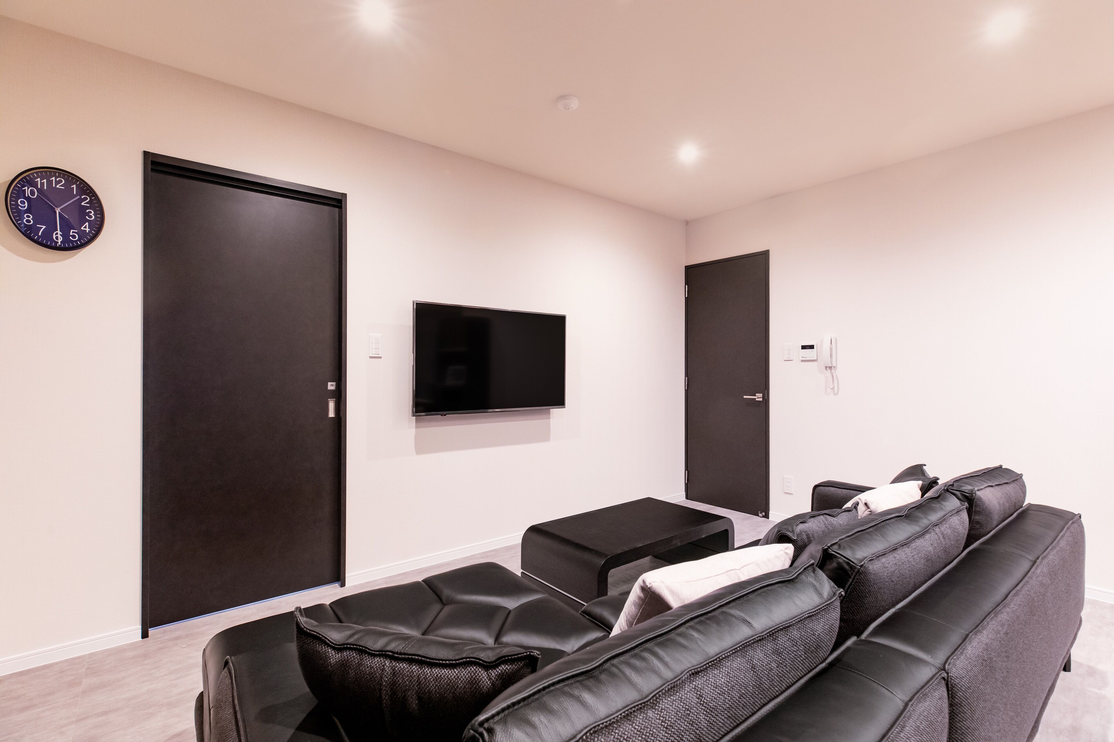 Un salon moderne avec un canapé en cuir noir, un téléviseur à écran plat fixé au mur et deux portes de couleur foncée.