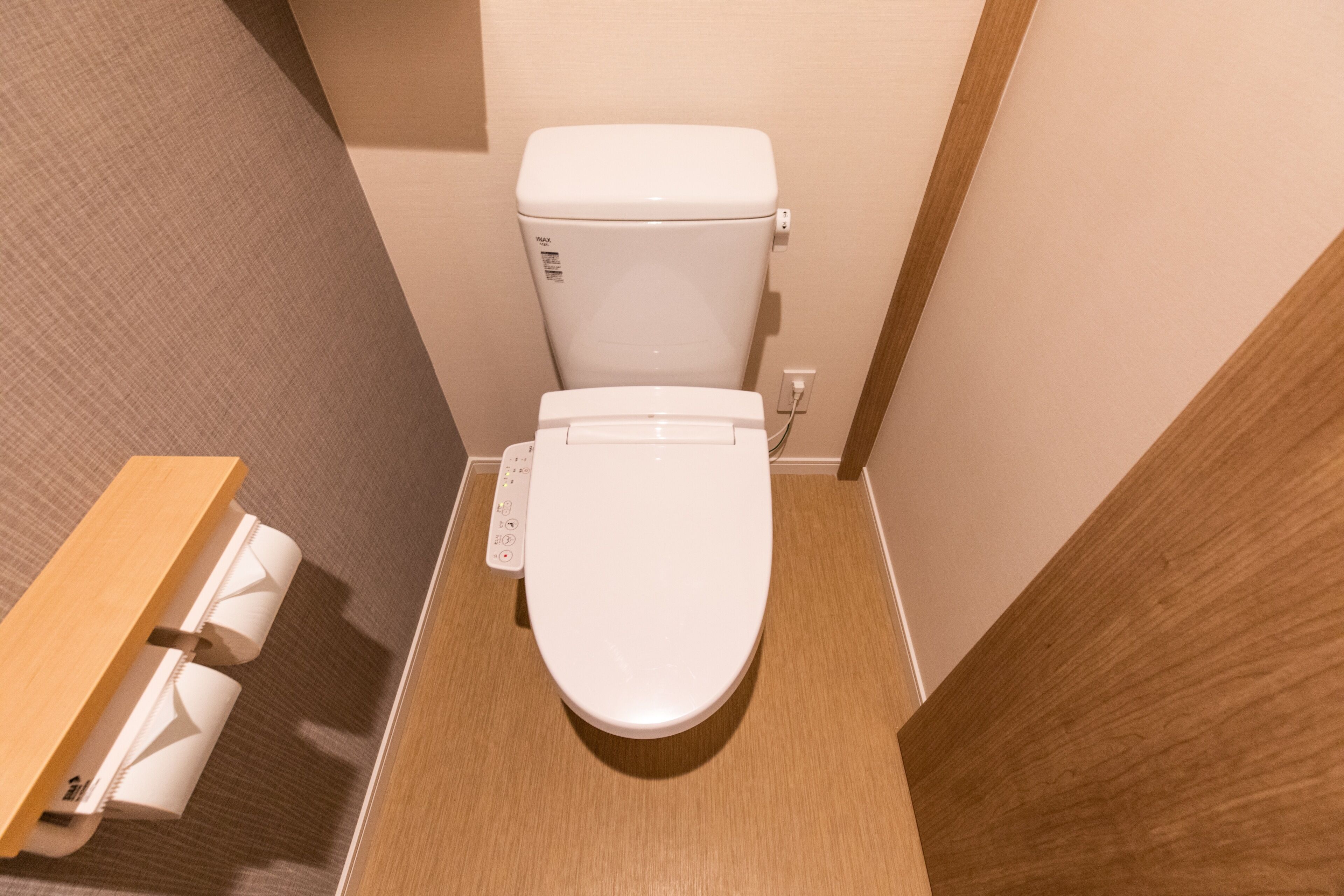 Et lille badeværelse med et hvidt toilet, en træhylde med toiletpapir og en ensfarvet væg.