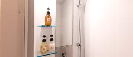 Casa de banho com chuveiro, prateleira de vidro com produtos de higiene e uma porta.