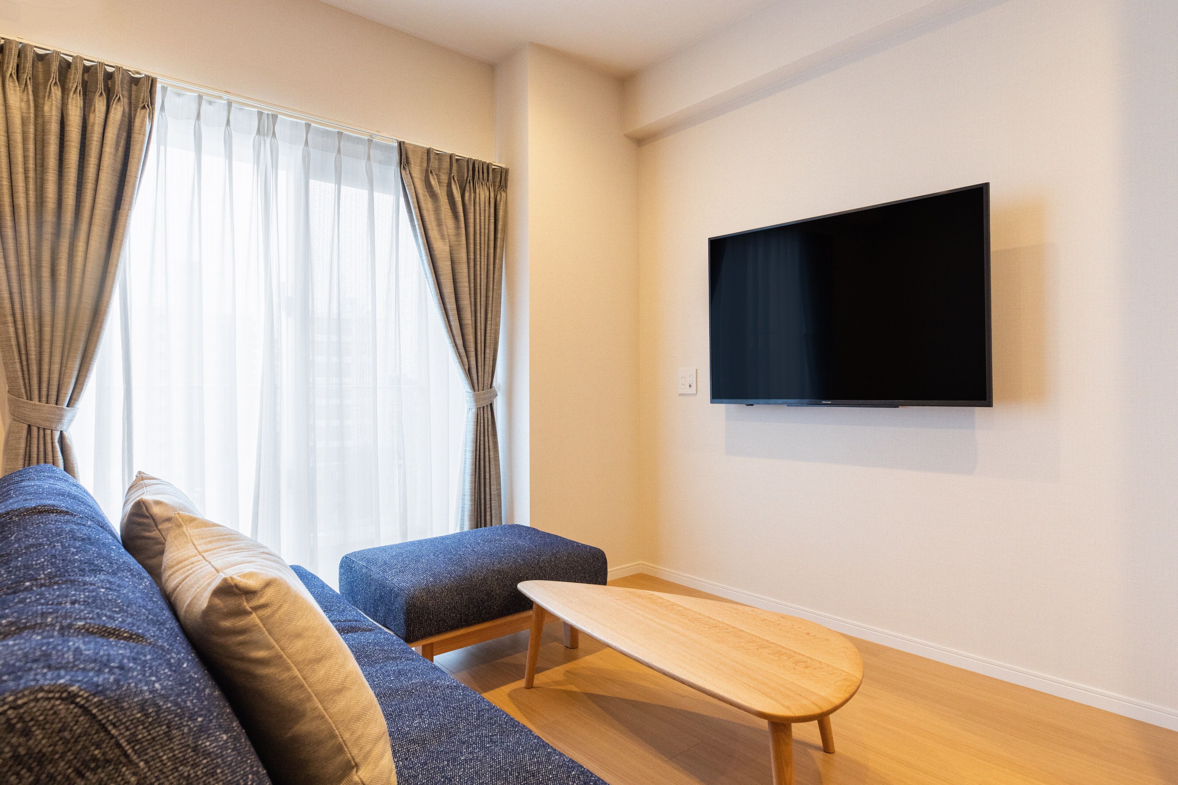 Een moderne woonkamer met een bank, poef en salontafel. Een flatscreen televisie is aan de muur bevestigd.