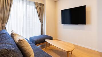Een moderne woonkamer met een bank, poef en salontafel. Een flatscreen televisie is aan de muur bevestigd.