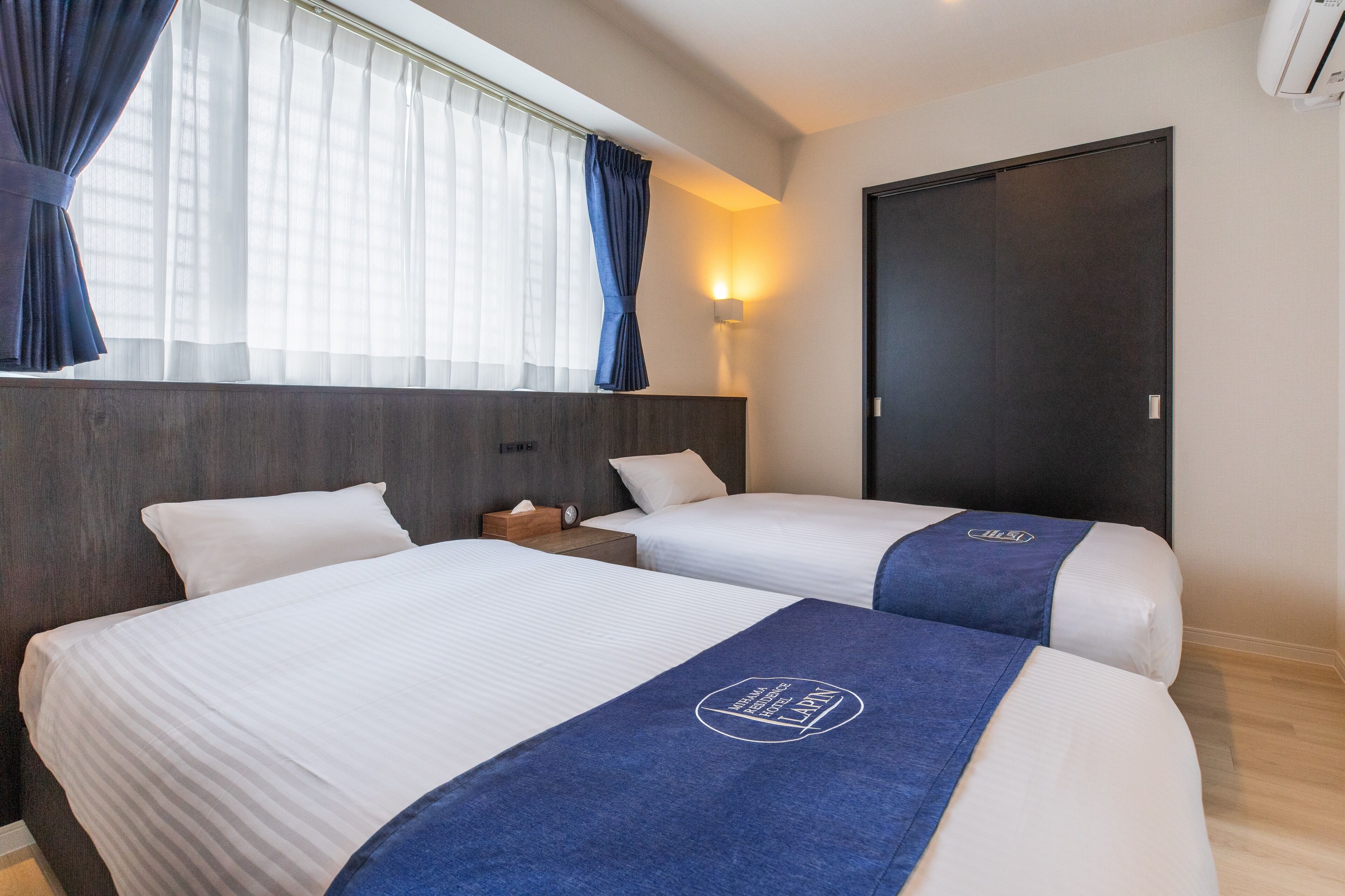 Une chambre d’hôtel avec deux lits, une tête de lit en bois, une table de chevet, une lampe et une fenêtre avec des rideaux.