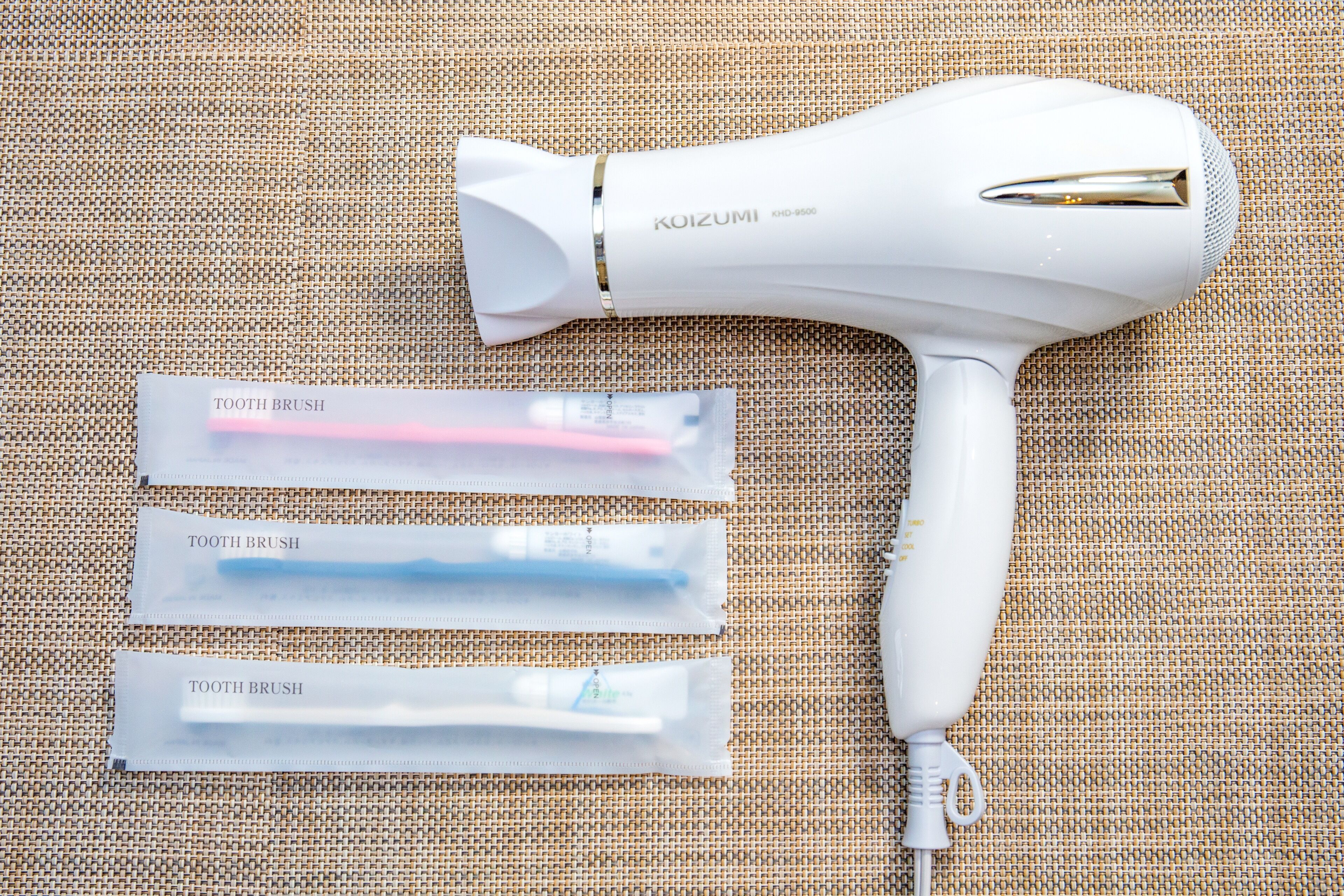 A white hair dryer with the brand name 'KOIZUMI' and three tooth brush packets labeled 'TOOTH BRUSH' on a textured surface.