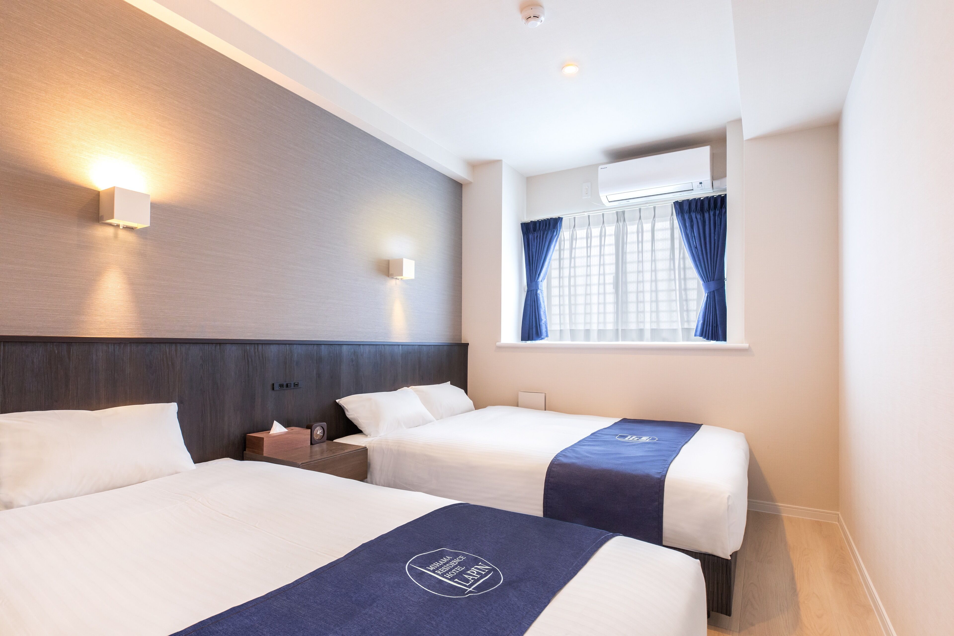 Quarto de hotel com duas camas, mesa de cabeceira, ar condicionado fixado na parede e janela com cortinas.