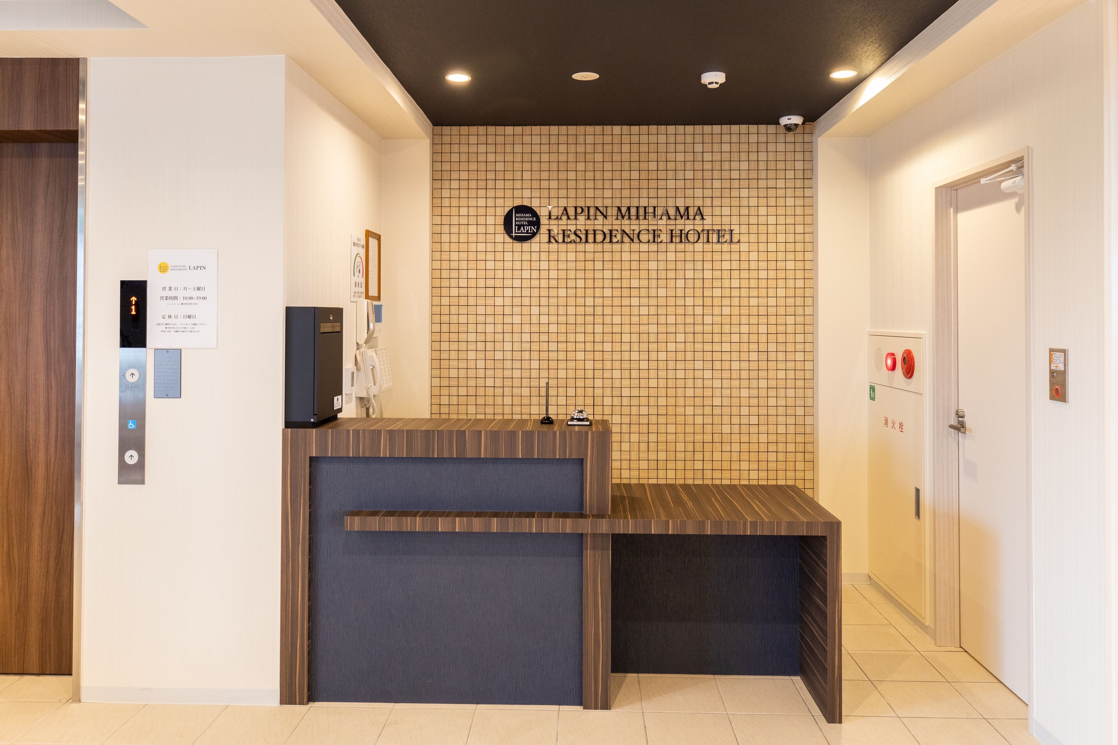 Mostrador de recepción en el Lapin Mihama Residence Hotel, con un muro de azulejos, una puerta de madera y un ascensor.