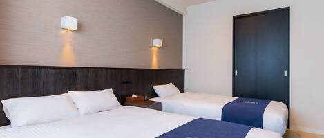 A hotel room with two beds, a dark wooden headboard, two wall-mounted lights, a dark door, and a small wooden nightstand with a lamp.