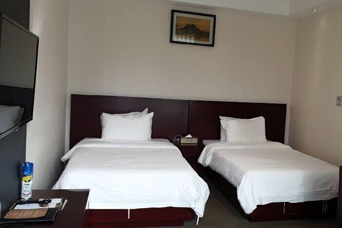A hotel room with two single beds, a dark-colored headboard, a small table with a bottle and papers, a chair, and a door.