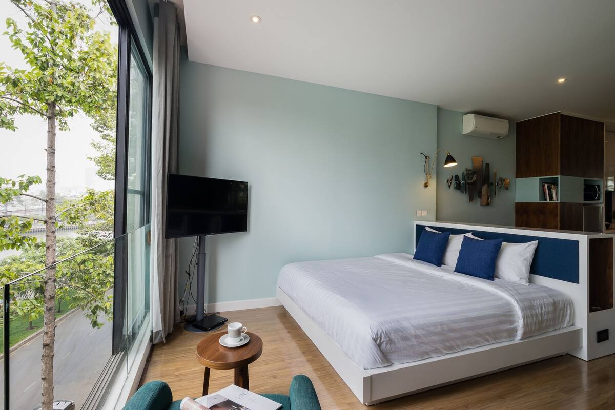 Ein modernes Hotelzimmer mit einem großen Bett, einem Fernseher, einem kleinen Tisch mit einer Tasse und Blick auf Bäume durch das Fenster.